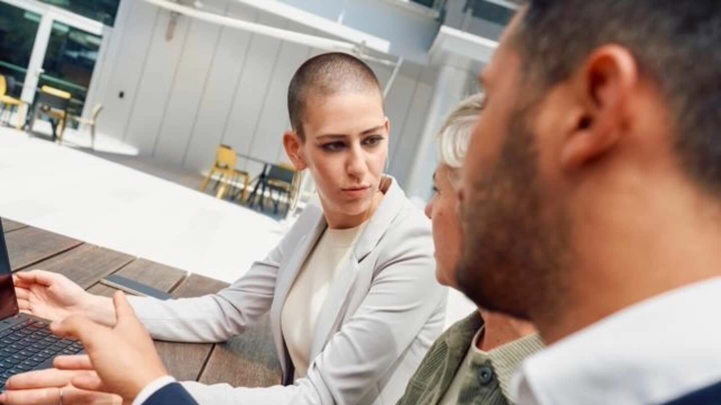 An image of a woman with a shaved head wearing neutral color business clothes talking to two people in the background.