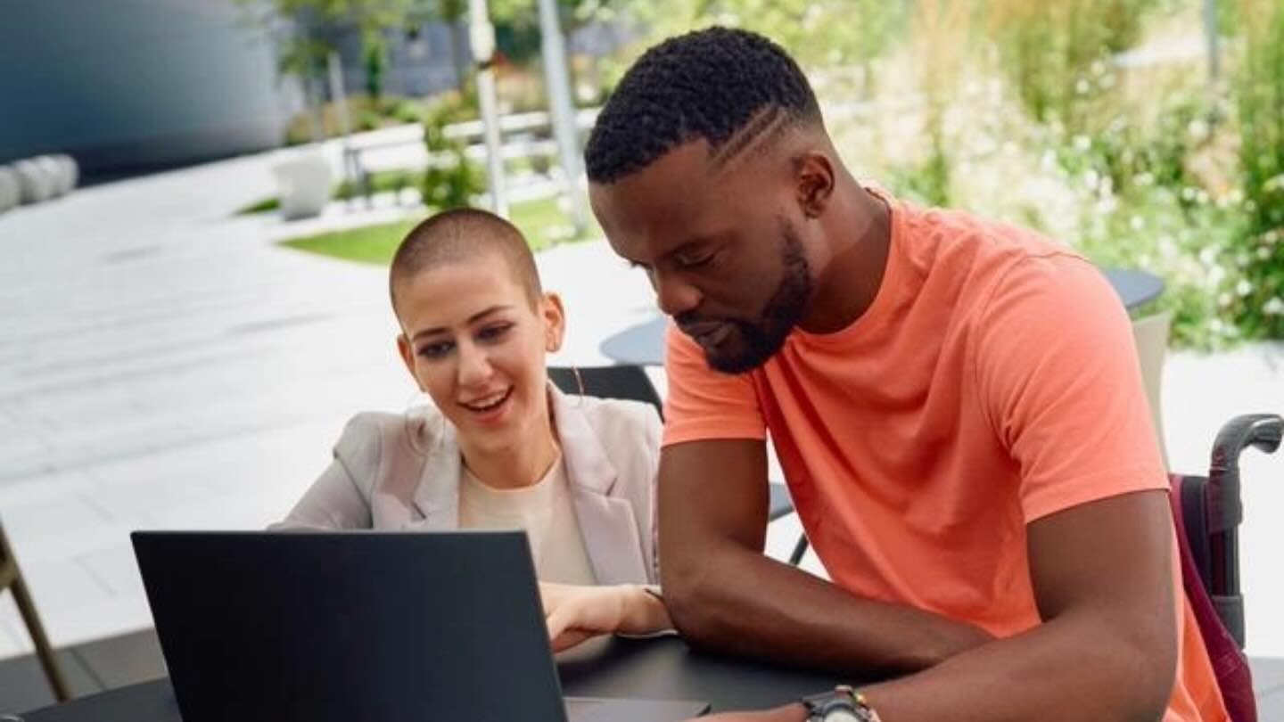 A photo of a white woman with a shaved head and a black man wearing a pink shirt sitting at a table looking at a computer screen